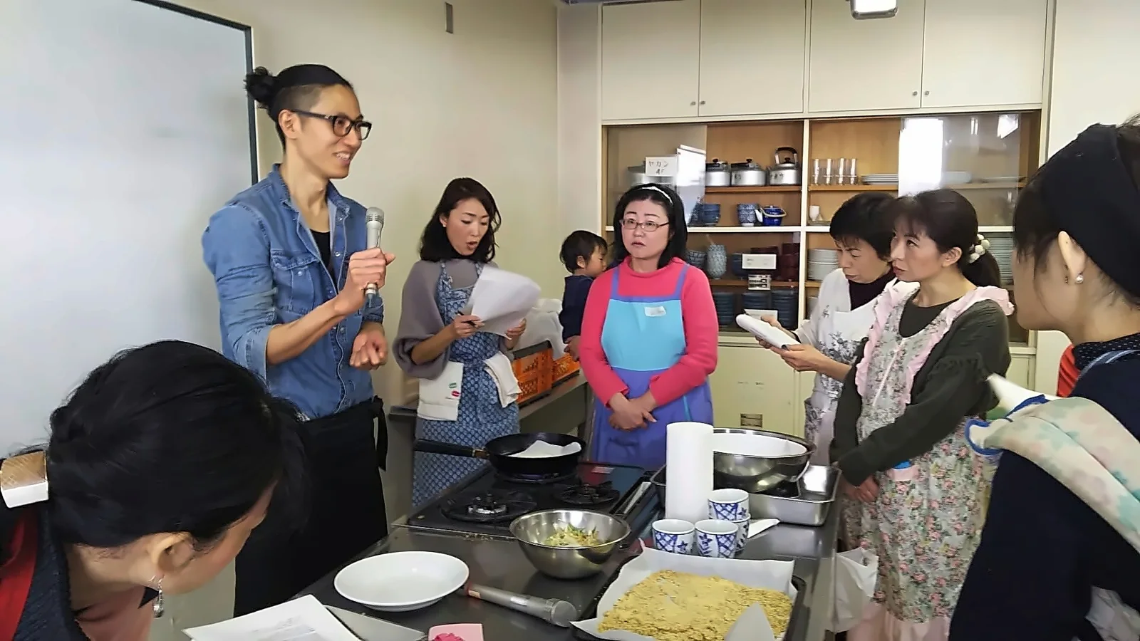 講師がマイクを持ちながら、受講生に囲まれて実演を交えて説明する料理教室の様子